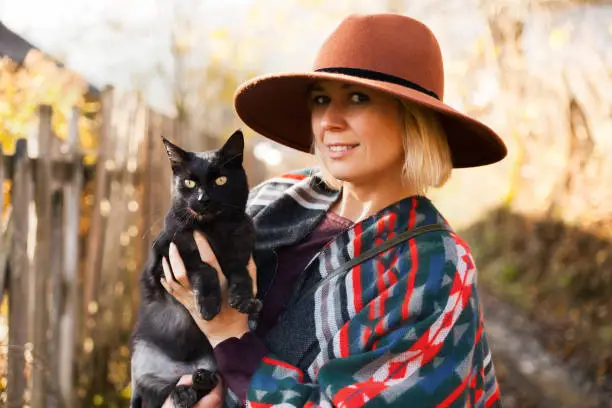 Black cat and stylish traveling woman wearing  authentic boho chic style poncho and hat near old wooden countryside buildings Woman have a fun in autumn time.Travel and wanderlust concept.