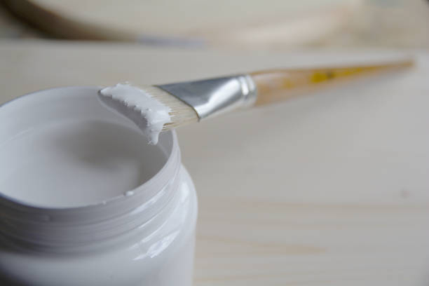 Plastic can with white acrylic primer and a bristle brush on a wooden panel. Preparing the surface for drawing.
