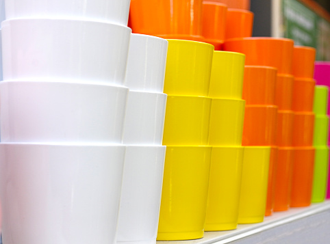 Many colored ceramic flower pots are stacked on a shelf in a garden equipment store. Yellow, white, orange, green and pink clay pots are sold for interior decoration. A large group of new gardening tools for planting
