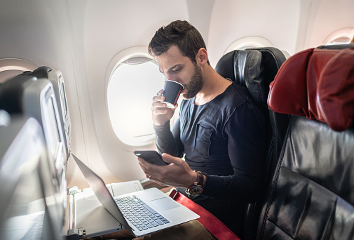 Man working in airplane using cellphone and drinking coffee