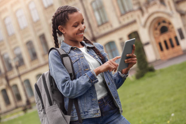 istock Young schoolgirl in jeans with backpack standing at school yard browsing igital tablet cheerful 1165006274