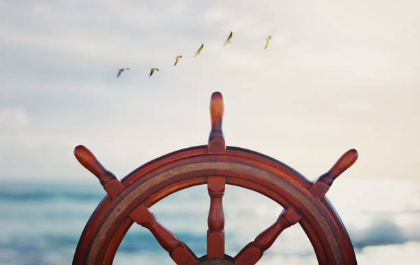 vintage nautical detail of a steering wheel of a ship in front of the sea at sunset vintage nautical detail of a steering wheel of a ship in front of the sea at sunset rudder stock pictures, royalty-free photos & images