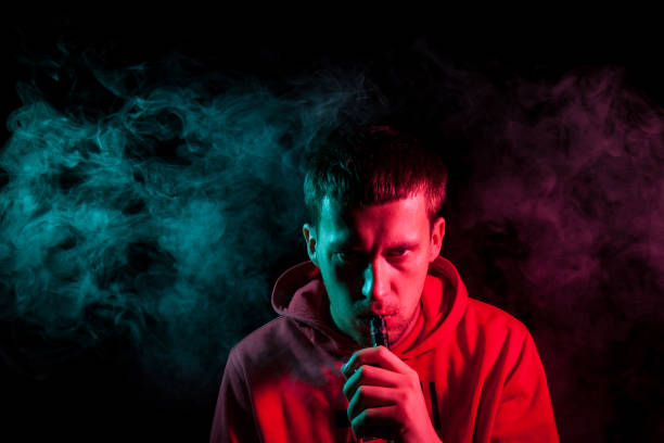istock A young brunette man holds in his hand and drags on an electronic cigarette with a vape while smoking and releases smoke to the sides, his face is highlighted with red and green light. 1165071834