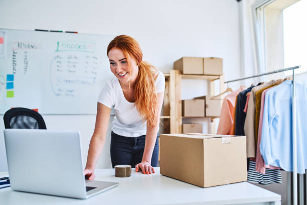 joven propietario de negocios en línea mirando laptop mientras prepara entregas para clientes - e commerce shopping women internet fotografías e imágenes de stock