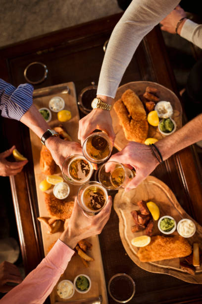 Cheers to Friendship Direct above images of unrecognisable people eating fish and chips on wooden plates. The males are doing a toast with their pints of beer. pub food stock pictures, royalty-free photos & images