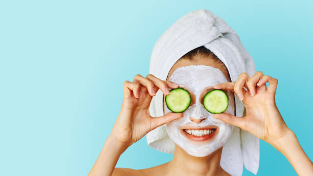 Beautiful young woman with facial mask on her face holding slices of cucumber Skin care and treatment, spa and cosmetology concept. Over blue background with copy space cucumber photos stock pictures, royalty-free photos & images