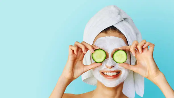 Photo of Beautiful young woman with facial mask on her face holding slices of cucumber