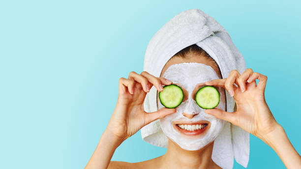 beautiful young woman with facial mask on her face holding slices of cucumber - facial mask spa treatment cucumber human face imagens e fotografias de stock