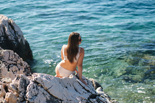 female sitting on the rock in the sea. Attractive young woman in beautiful swimsuit by the sea. Splash from the waves.