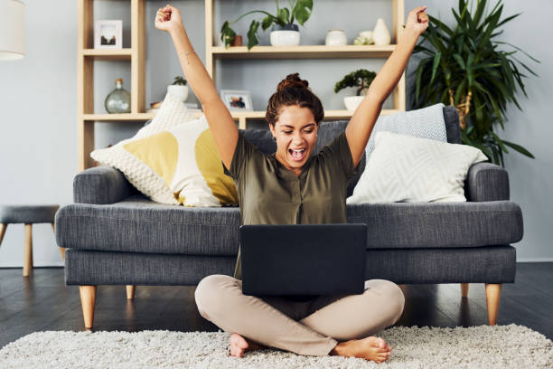 Entering all those competitions finally paid off Shot of a young woman cheering while using a laptop on the living room floor at home ecstasy stock pictures, royalty-free photos & images
