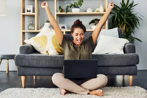 Shot of a young woman cheering while using a laptop on the living room floor at home