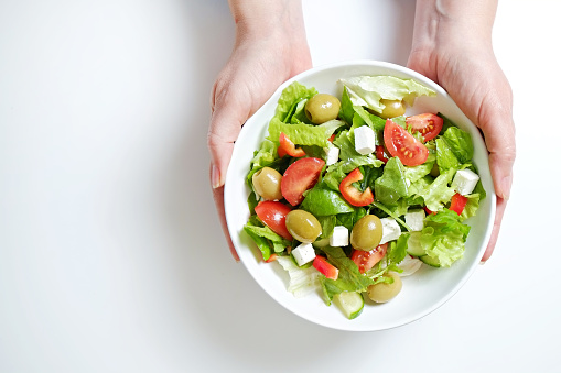 Traditional greek salad with fresh ingredients, feta cheese, olives, red tomatoes, cucumbers and greens in ceramic bowl, isolated on white background. Top view, close up, copy space.