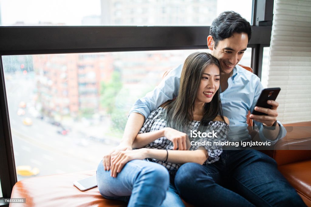 Joyful couple using smart phone together at home Chinese woman and mixed race man sitting on sofa at apartment and using smart phones together Couple - Relationship Stock Photo