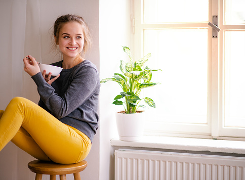 A happy young female student sitting by window, eating. Copy space.