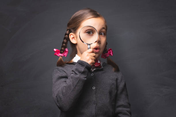 Child Girl Student Looking Through Magnifying Glass on Blackboard Backdrop with Available Copy Space. Back to School Concept.