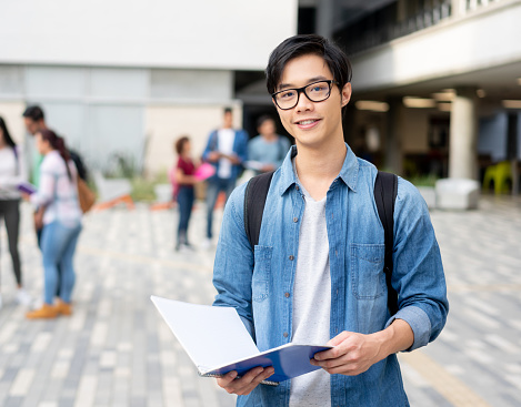 Young smart male collage student in casual clothings and backpack smiling, standing outdoor with campus building in the background and holding books while looking at camera ready for study. Education concept.