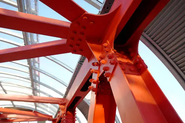 Photo of Red iron beams bolted on. Angular connection of several iron beams in one place. Pedestrian crossing with iron construction and glass ceiling.