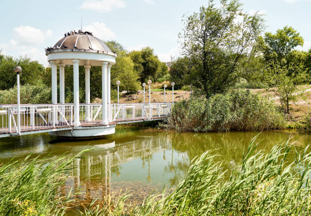 Pavilion in beautiful public garden park. White Gazebo forest view. Outdoor arbor with forged bridge, pond and background of a summer landscape. lonely arbour near river. Fine garden with green grass.