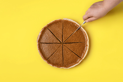 Slicing a pie concept. Woman hand cutting in slices a pumpkin pie on a yellow background. Top view of delicious traditional Thanksgiving dessert.