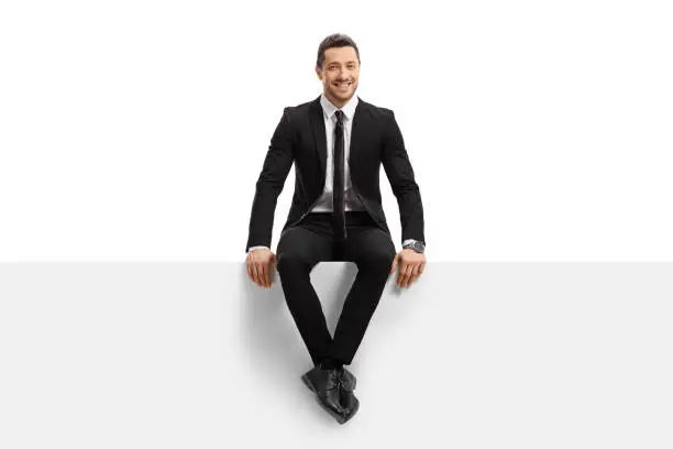 Full length portrait of a young man in a suit sitting on a panel and smiling isolated on white background