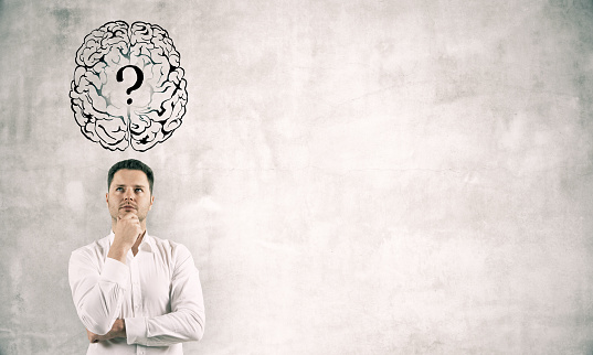 Thoughtful young businessman standing on white concrete wall background with brain and question mark sketch. Brainstorm and confusion concept