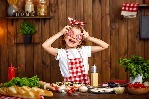 Adorable little female chef wearing checkered apron and headband cooking pizza and making face with salami slices instead eyes and opened mouth on stylish wooden kitchen