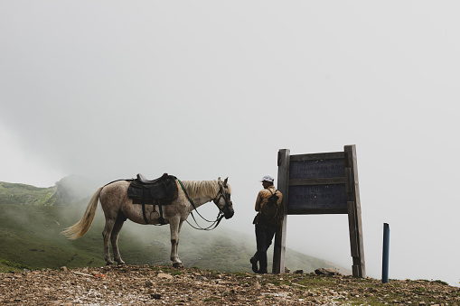a man stands leaning on a road sign next to his horse.it's like he's resting after a long journey.