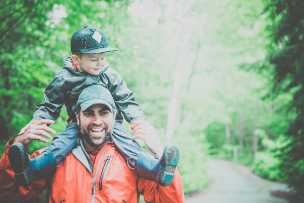 A father is carrying his son on his shoulders in the forest. It is a cloudy summer day but they are both smiling and having fun.