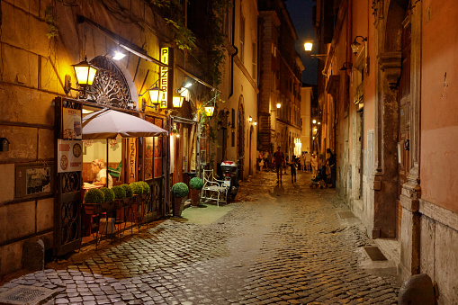 Pizzeria restaurant Le Copelle in Via Delle Coppelle Alley in old town of Rome nightime. A woman is having her dinner while some people are walking in the alley. Rome is the capital city of Italy.
