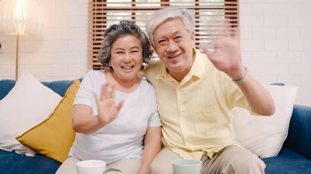 Asian elderly couple using smartphone video conference with grandchild while lying on sofa in living room at home. Enjoying time lifestyle senior family at home concept. Portrait looking at camera.