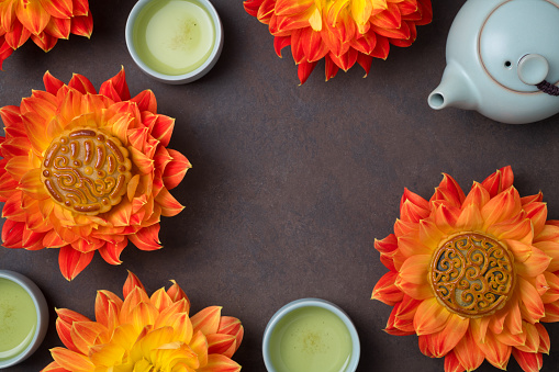 Mooncakes in a fresh orange dahlia flowers, a blue teapot and cups of green tea on a brown background. Chinese mid-autumn festival food. Top view.