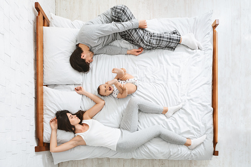 Joint sleep with kids. Exhausted parents sleeping on sides of bed, active baby playing in middle, above view