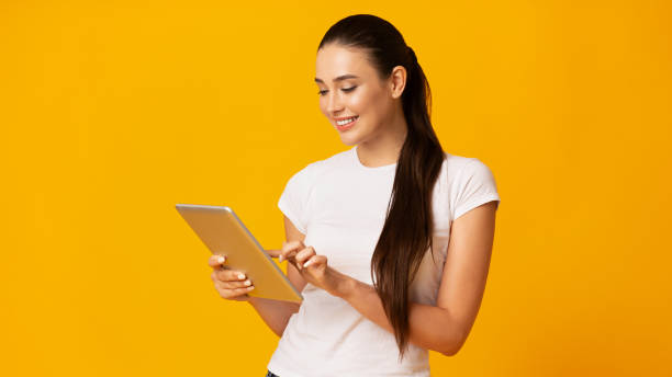 fille de sourire utilisant l'ordinateur de tablette sur le fond jaune - digital tablet women enjoyment happiness photos et images de collection