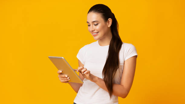 smiling girl using tablet computer on yellow background - digital tablet women enjoyment happiness imagens e fotografias de stock