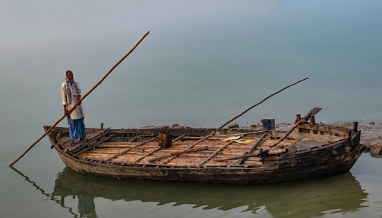 Middle Aged Indian Man rowing his wooden boat alone, towards river Ganges bank, in freezing winters of North India. Visibility is very low due to fog in winters. He is wearing kurta, lungi and muffler