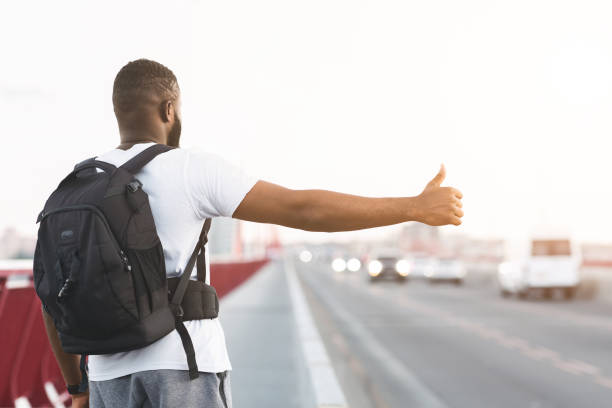 African american young man with backpack hitchhiking Young Black Guy Is Hitchhiking On The Bridge Road, carrying backpack, rear view hitchhiking stock pictures, royalty-free photos & images