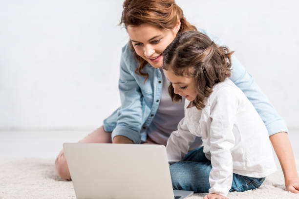 istock Cheerful mother and daughter sitting on floor and using laptop on white 1165269374