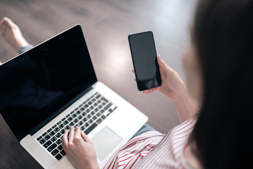 Young woman sits on a floor and uses a laptop and smartphone. Black blank screen of laptop and smartphone with real reflections for your mockup or design.