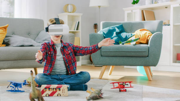 istock Smart Little Boy Wearing Virtual Reality Headset Uses Hand Gestures to Control Augmented Reality Gameplay. He's Sitting on a Carpet in His Living Room. Happy Child Uses Futuristic AR Glasses at Home. 1165279282