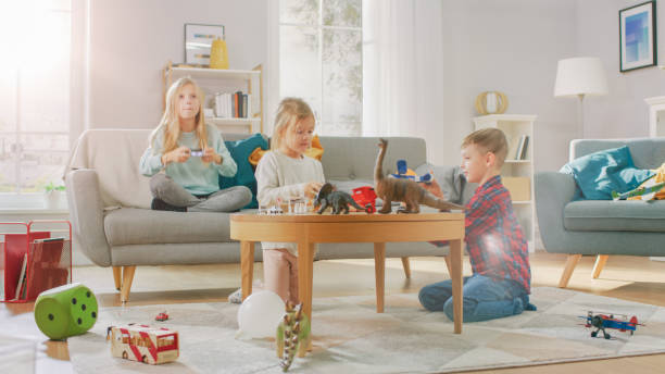 istock At Home: Cute Girl Playing in Video Game Console, Using Joystick Controller, Her Younger Brothe Plays with Toy Airplane and Youngest Sister Plays with Toy Dinosaurs. Happy Children Playing in the Sunny Living Room. 1165279370