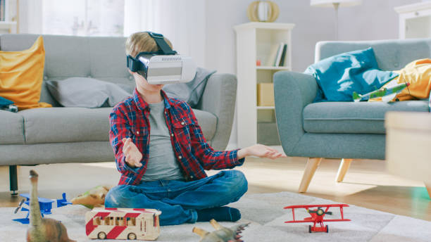 istock Smart Little Boy Wearing Virtual Reality Headset Uses Hand Gestures to Control Augmented Reality Gameplay. He's Sitting on a Carpet in His Living Room. Happy Child Uses Futuristic AR Glasses at Home. 1165279373