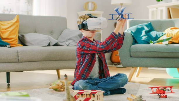 istock Smart Little Boy Wearing Virtual Reality Headset Plays with Toy Airplane while Sitting on a Carpet in His Living Room. Happy Child Uses Futuristic AR Glasses at Home. 1165279476