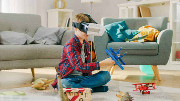 istock Smart Little Boy Wearing Virtual Reality Headset Plays with Toy Airplane while Sitting on a Carpet in His Living Room. Happy Child Uses Futuristic AR Glasses at Home. 1165279521