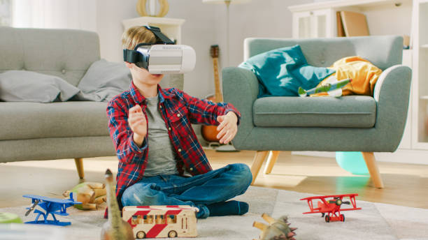 istock Smart Little Boy Wearing Virtual Reality Headset Uses Hand Gestures to Control Augmented Reality Gameplay. He's Sitting on a Carpet in His Living Room. Happy Child Uses Futuristic AR Glasses at Home. 1165279538