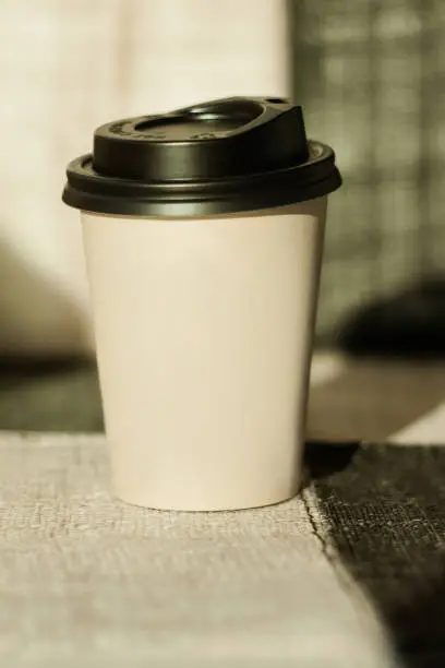 Kraft paper coffee cup with black lid from street shop with hot aroma drink near the window. Lifestyle concept, take away food. Copy space, front view.