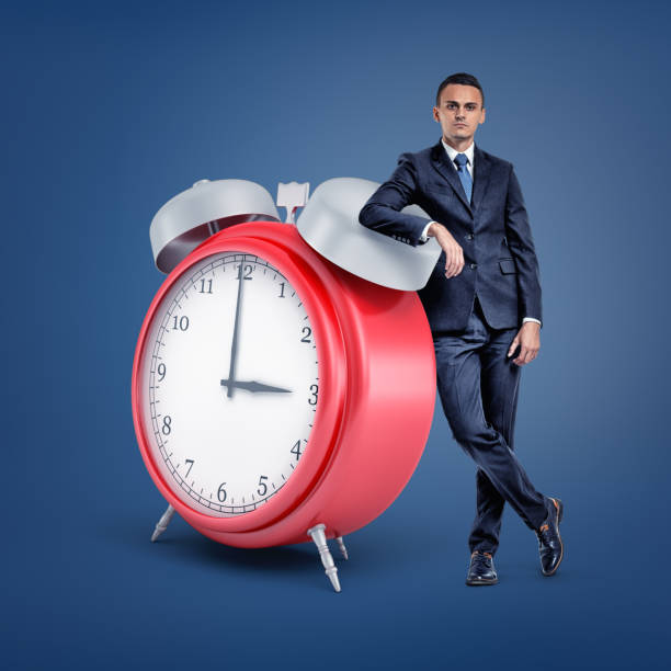 istock A small businessman stands looking at front and leaning on a huge red alarm clock. 1165289881