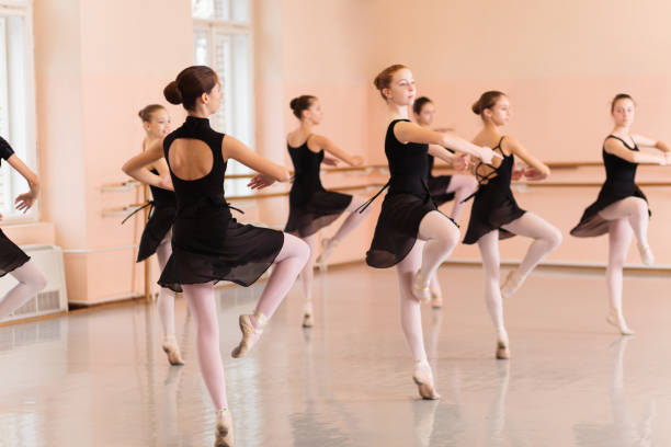 grupo mediano de adolescentes con vestidos negros practicando movimientos de ballet en un gran estudio de baile - estudio de ballet fotografías e imágenes de stock