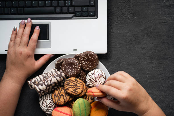 Woman eating cookies and coffee at workplace Unhealthy snack at work time. Compulsive indulgence, overeating, stress, high calorie, fattening junk food, weight gain. Woman eating cookies at workplace over eating stock pictures, royalty-free photos & images