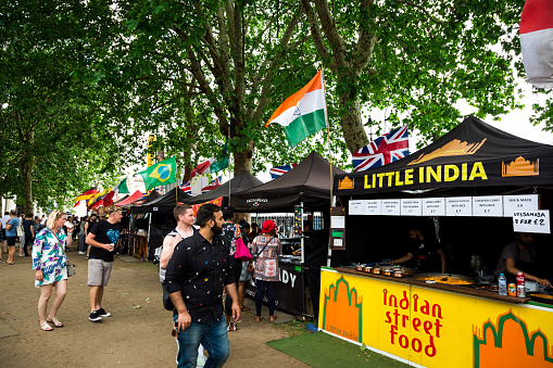 London, UK - 30 June, 2019: color image depicting crowds of people perusing and sampling the food from a range of different stalls at an outdoor food market in London, UK. The range of different cuisine on offer includes Greek, Indian, Brazilian and Mexican. Each of the food stalls has its country's flag flying proudly above it.