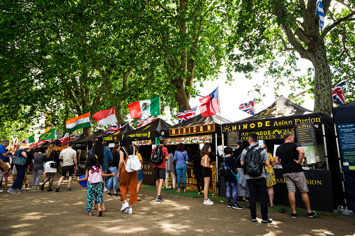 London, UK - 30 June, 2019: color image depicting crowds of people perusing and sampling the food from a range of different stalls at an outdoor food market in London, UK. The range of different cuisine on offer includes Greek, Indian, Brazilian and Mexican. Each of the food stalls has its country's flag flying proudly above it.